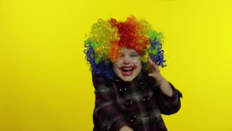 la niña payasa con la peluca colorida se esconde detrás de sus manos y muestra caras divertidas. halloween
