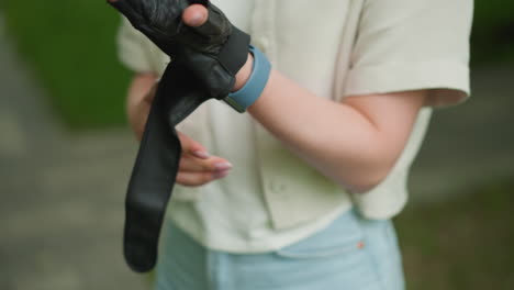 close-up of young woman putting on black gloves with focus on her hands and smartwatch on right wrist, background features blurred greenery and hints of outdoor path