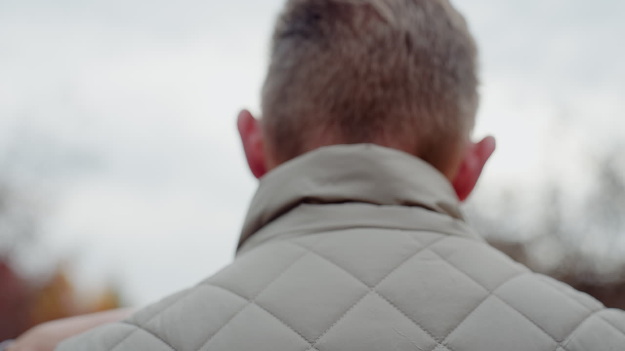 Back view of man in summer jacket being embraced by woman with pink painted nails resting hands on his shoulders as they share intimate hug outdoors under soft daylight with affection and connection