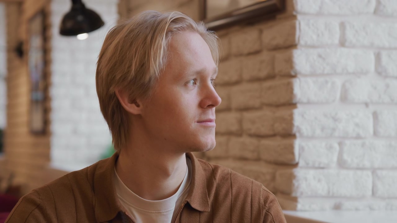 Close up of young man with short blonde hair wearing brown zip up shirt and white t shirt seated in indoor setting. subject remains stationary against soft lighting from hanging lamps