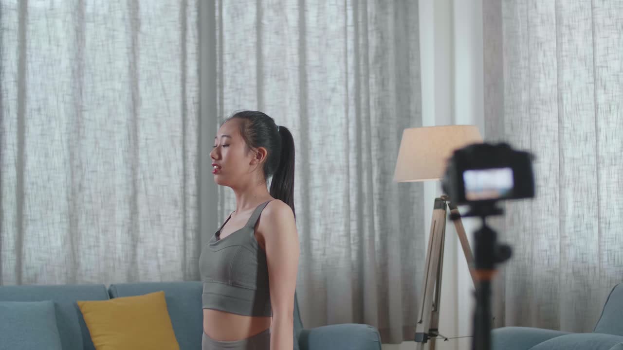 스포츠 옷을 입은 젊은 아시아 트레이너 여성이 카메라에 대화하고 집에서 운동을 가르치는 것을 녹음하는 동안 높은 지 자세로 요가를합니다.