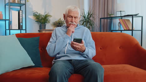 Senior-old-grandfather-sitting-on-sofa,-using-smartphone-share-messages-on-social-media-application