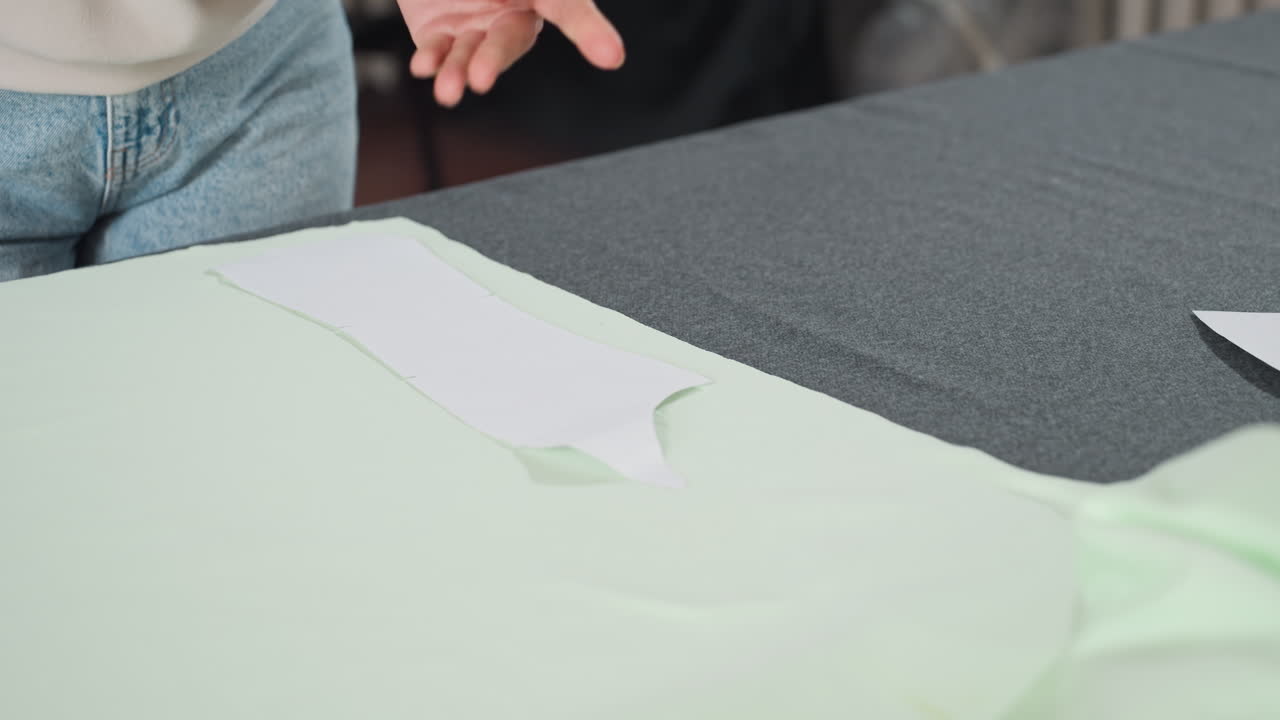 Fashion designer lays out paper cutting pattern precisely over spread light green fabric on workspace table in bright sewing studio, preparing fabric arrangement for professional garment making