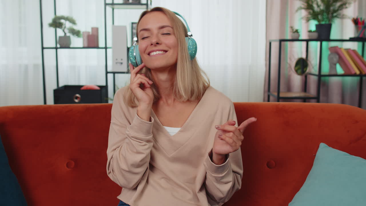무선 헤드폰을 입은 행복한 젊은 여성이 집에서 소파에 앉아 좋아하는 음악을 듣고 휴식을 취하고 있습니다.