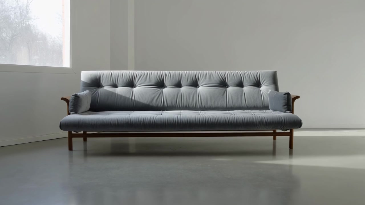 Tre-personers mid-century moderne sofa i afdæmpet blågråt stof står op ad en lysegrå væg, mens vindueslys skifter hen over de tuftede puder, hvilket skaber bløde højdepunkter, skygger og en rolig showroom-stemning.