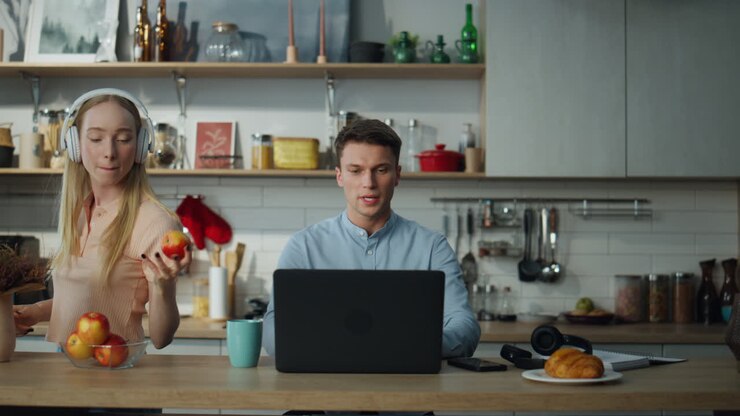 Empresario realizando negociaciones en línea en la cocina, esposa bailando con auriculares.