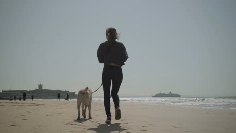 rear view of young girl running with dog on sandy seashore.