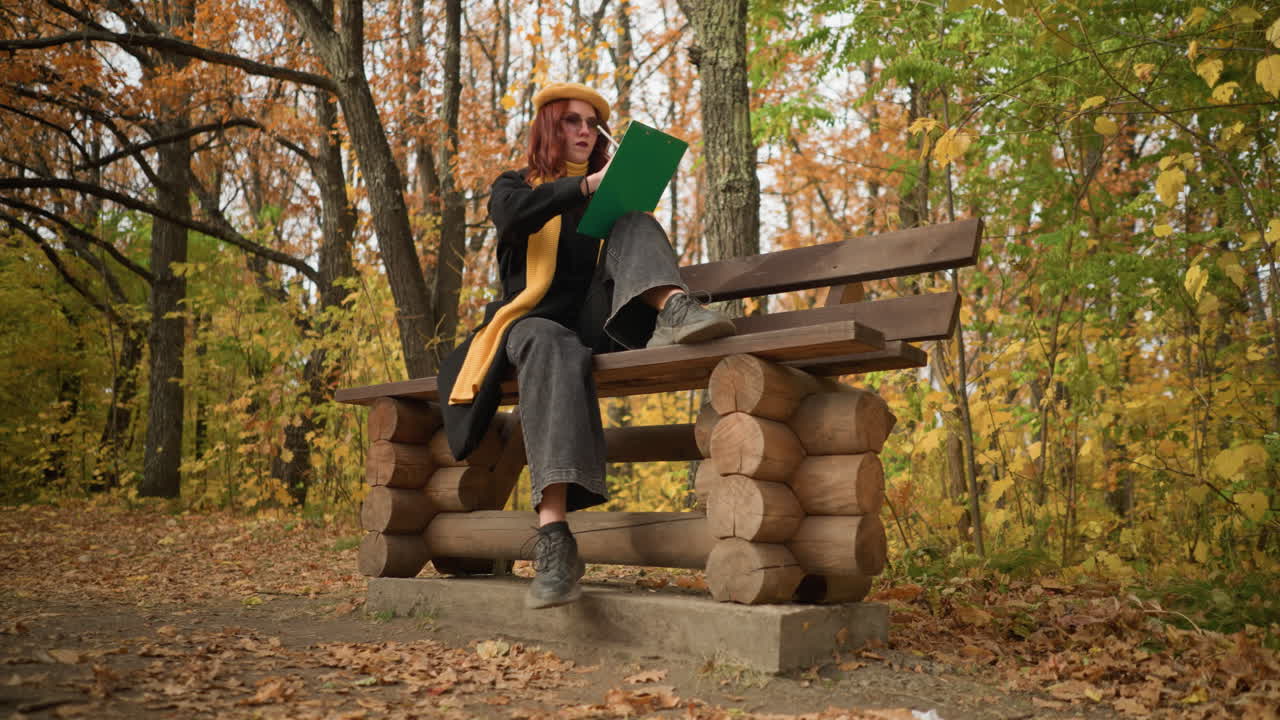 Artista elegante concentrato sullo schizzo mentre è seduto su una panchina di legno del parco in un sereno paesaggio autunnale, con una gamba sulla panchina e una a terra, circondato da foglie dorate e natura pacifica.