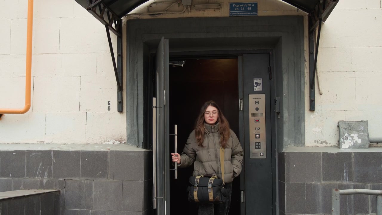 Mulher caucasiana saindo da entrada de um edifício urbano, apoiada em degraus de pedra e esperando pela campainha, momentos de passageira pensativa, estudante, musicista e entregadora se alternam enquanto ela verifica o telefone e ajusta a jaqueta