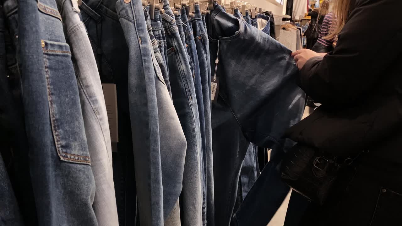 The woman looks through and touches the pants on the hangers in the clothing store. Pants offer in shops