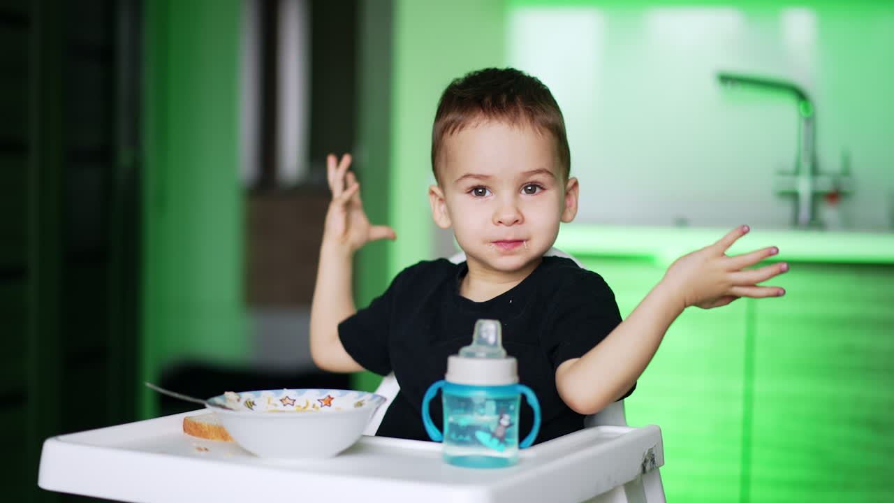 Cute baby drinking water from a bottle sitting at high chair. Mom cleans kid's mouth with a napkin. Blurred backdrop.