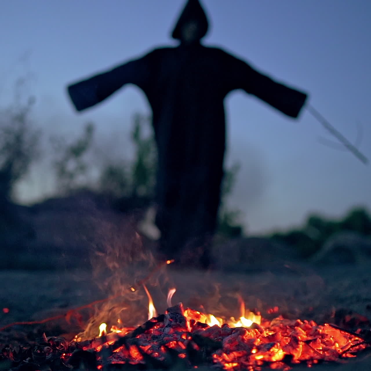 Embers of firewood and black scary silhouette. Mysterious figure in dark costume withwooden stick standing behind the smoldering logs of fire.