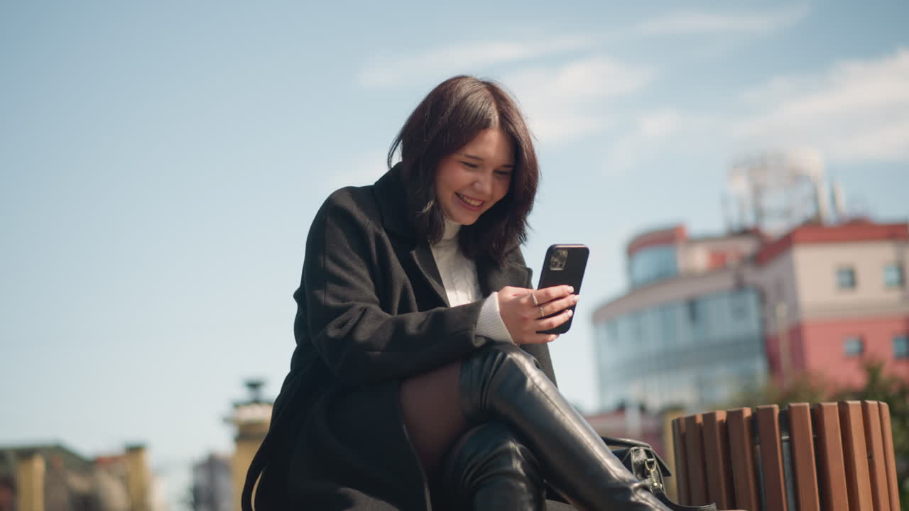 una joven blanca alegre sonriendo mientras se centra en su teléfono inteligente en un parque con las piernas cruzadas, un fondo borroso con un edificio y hierba verde, disfrutando de su momento al aire libre