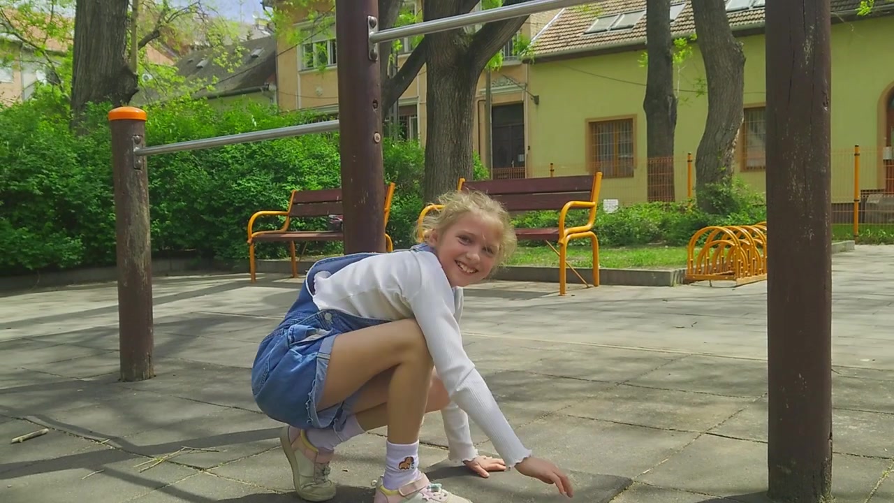 A medium shot captures a young girl in denim overalls performing an inverted handstand while hooked onto a metal pull-up bar at an outdoor park, preparing to dismount safely onto her feet.