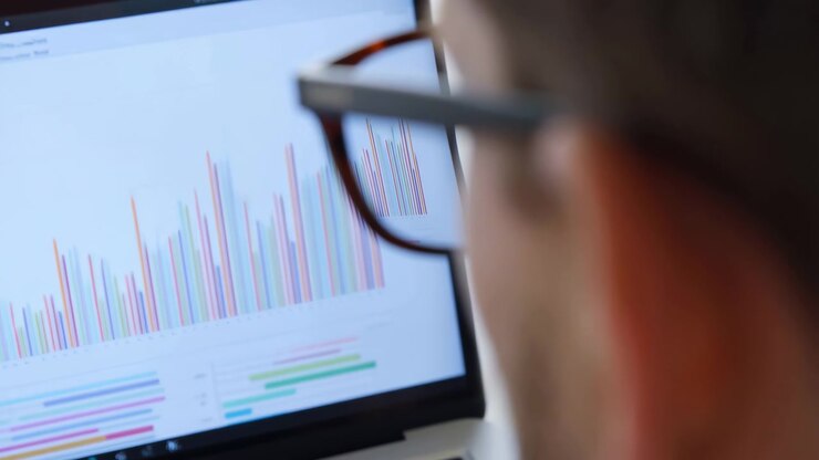 Vista ravvicinata da sopra la spalla di una persona con gli occhiali che esamina attentamente lo schermo di un laptop, pieno di barre verticali raggruppate, grafici orizzontali più piccoli e analisi aziendali pulite in un luminoso ambiente d'ufficio.