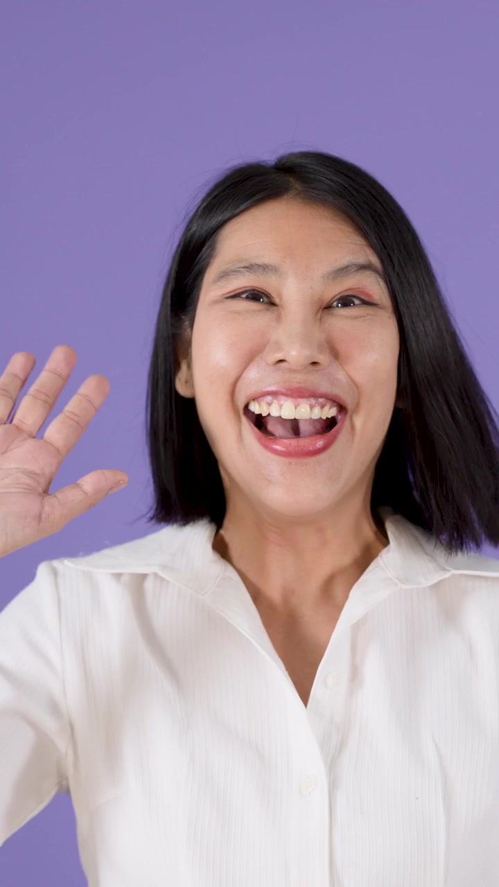 Una donna asiatica allegra con i capelli neri corti sorride e saluta su uno sfondo viola da studio