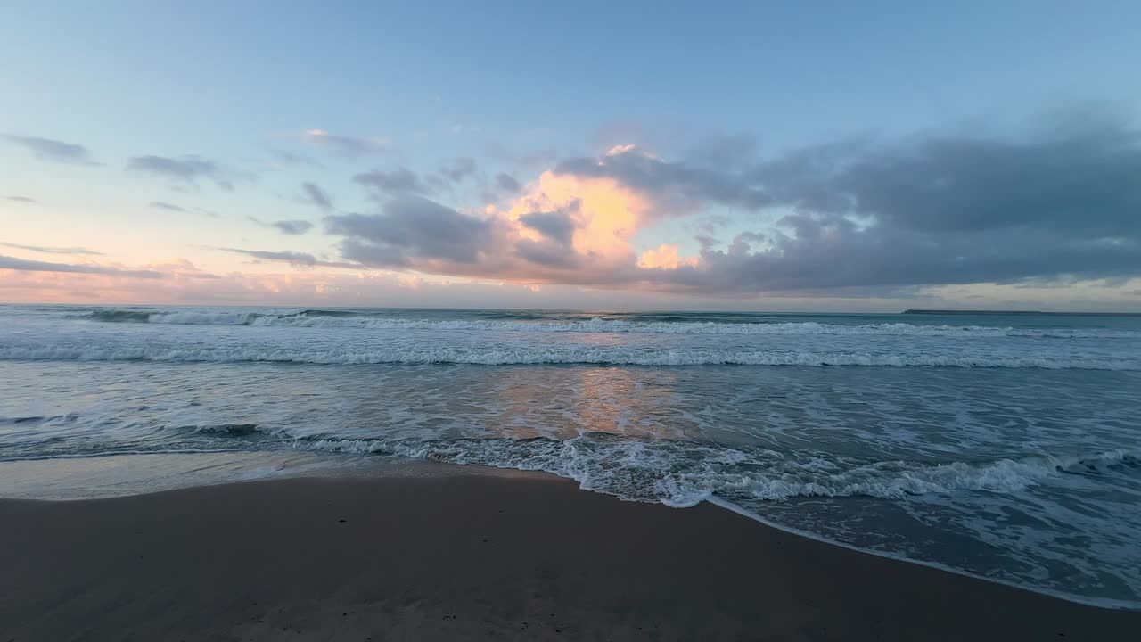 A wide view of a peaceful beach at sunset, where gentle waves roll to the shore under a softly clouded sky