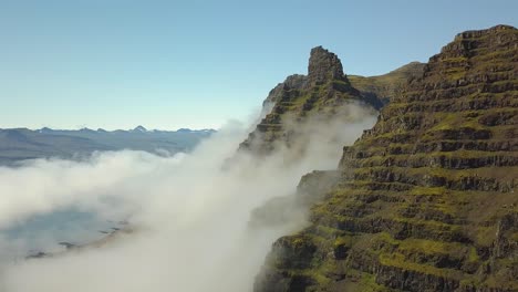 Aerial-rising-over-verdant-steep-hill-summit,-clouds-covering-mountains-in-background,-in-Djúpivogur-small-town-near-Iceland-fiords-at-daytime