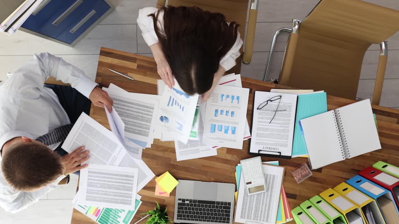 Overhead view of a business meeting at a wooden desk with documents, charts, and office supplies