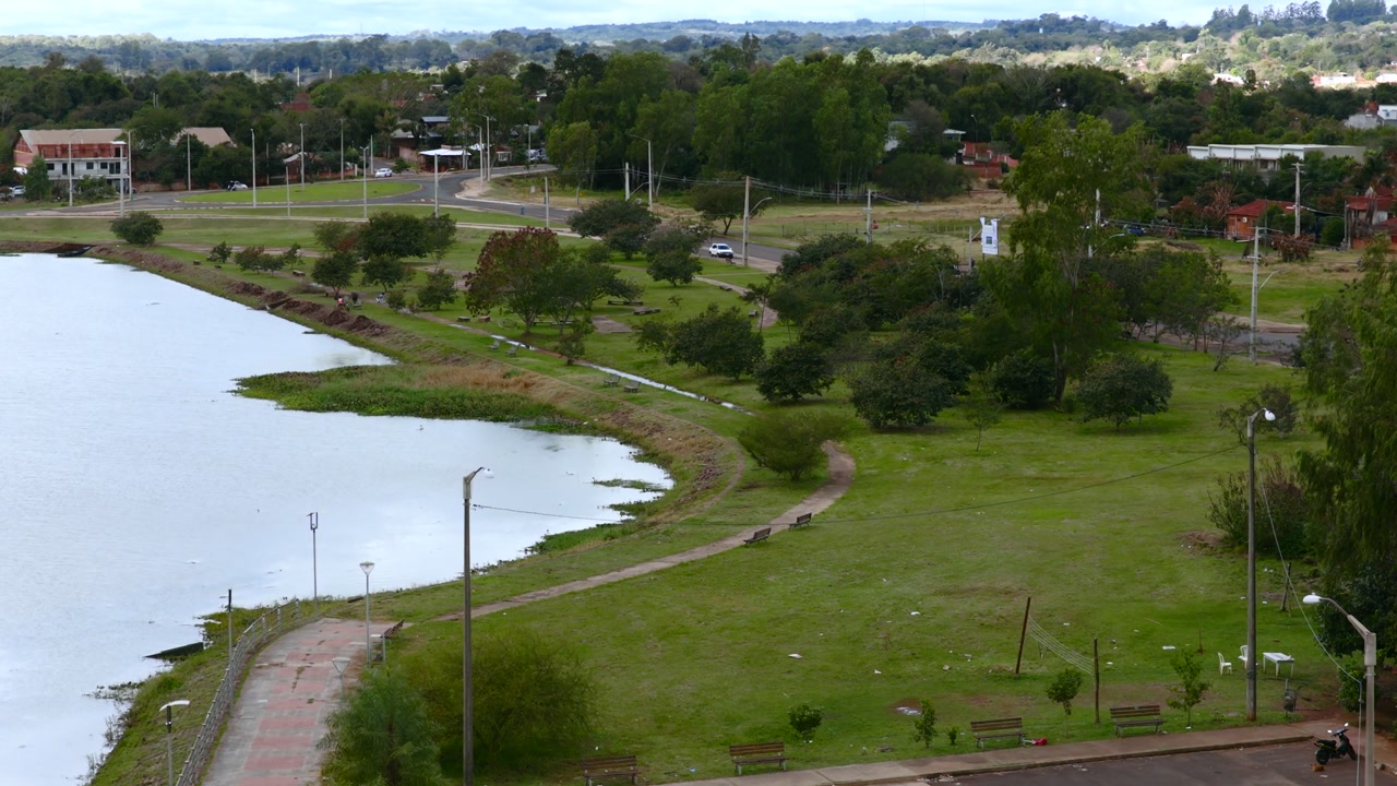 Captivating aerial footage of Mirador Cambyreta, showcasing a tranquil lakeside park with lush greenery and a scenic path. The serene atmosphere is enhanced by the lake's reflective waters.