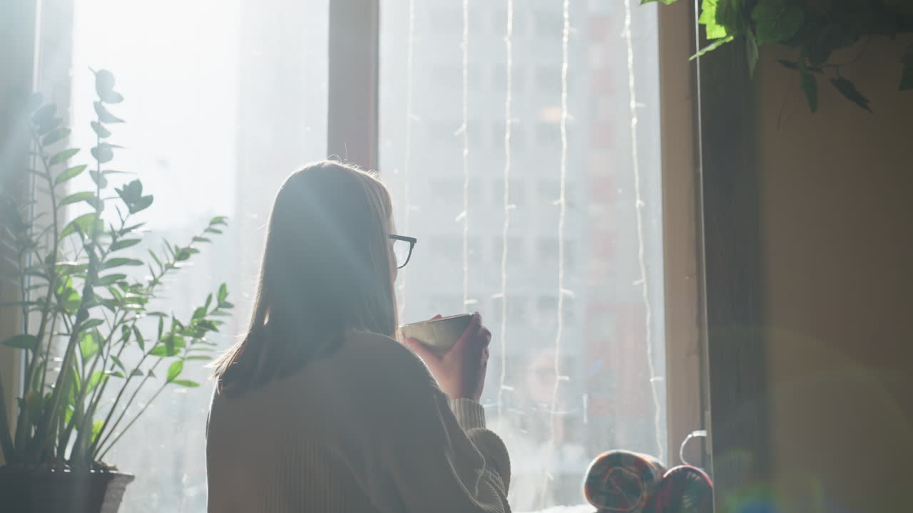 Sunlight glows around woman sipping coffee near window with plant and rolled cloths on table, city buildings appear through window as passerby moves in background