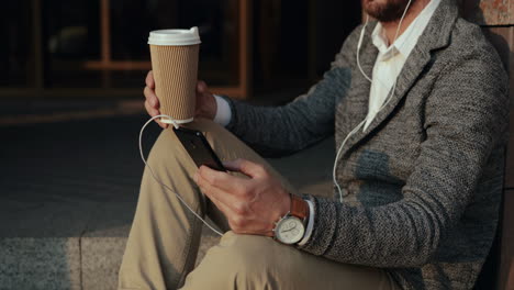 close up of a relaxed businessman drinking coffee and listening music on the smartphone via earphones while sitting on steps of a building in the city