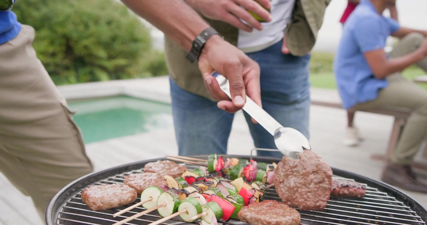 Hombres diversos observan a un hombre que revisa hamburguesas, sosteniendo pinzas para presionar y voltear brochetas en la parrilla junto a la piscina.