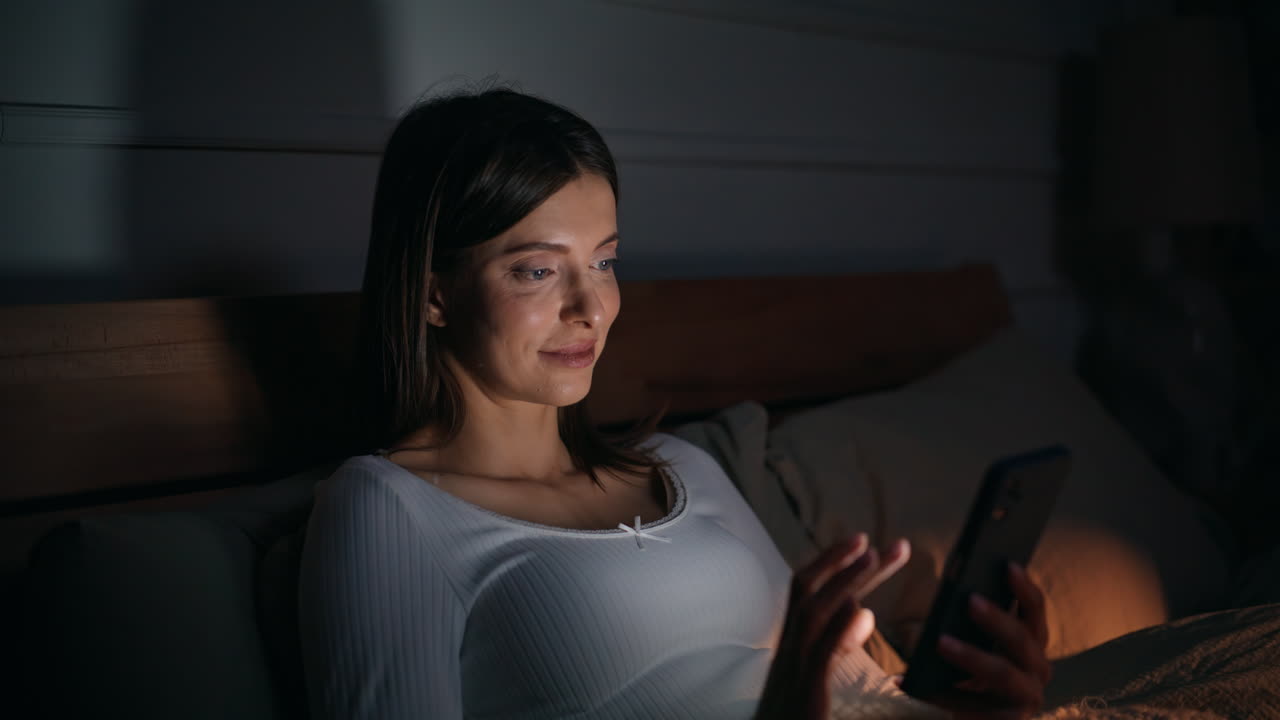 Lady scrolling phone dark bedroom relaxing on comfortable bed pillows closeup