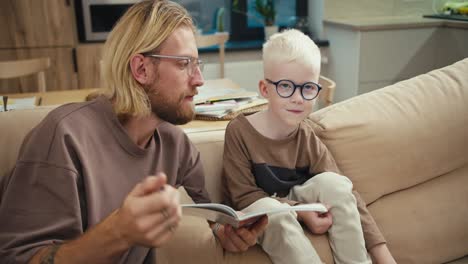 padre feliz, un hombre rubio con gafas y barba explica a su hijo pequeño, un niño albino con el color del cabello blanco en gafas azules, cómo leer y completar una tarea en la escuela primaria durante la tarea en casa en la cocina