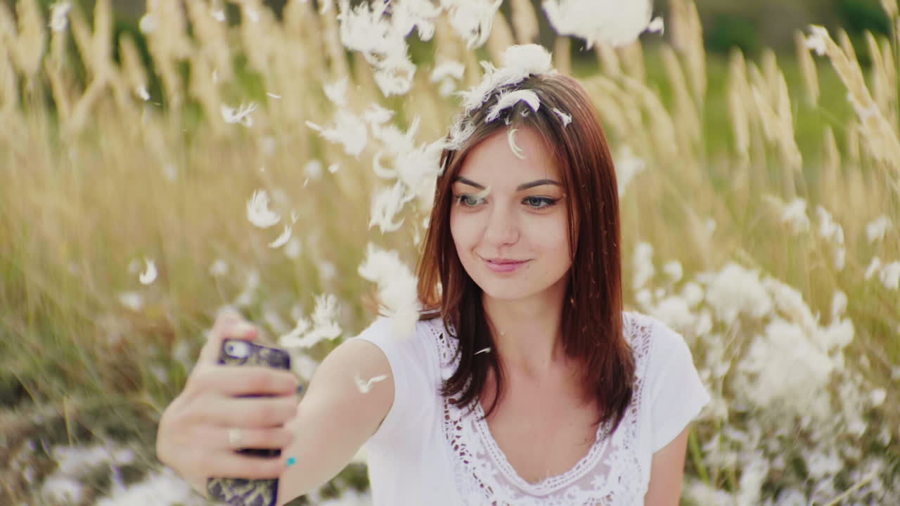 한 여성이 창의적인 사진 촬영에서 셀카를 만듭니다. 모든 것이 깃털 슬로우 모션 비디오로 흩어져 있습니다.