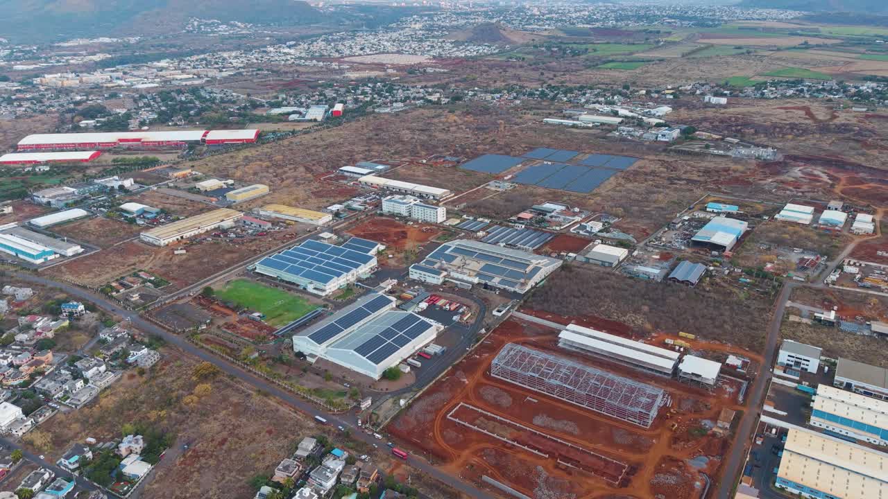 Flyover of Mauritius showing an extensive industrial area with large buildings covered in solar panels surrounded by dry terrain. Roads and infrastructure connect the factories
