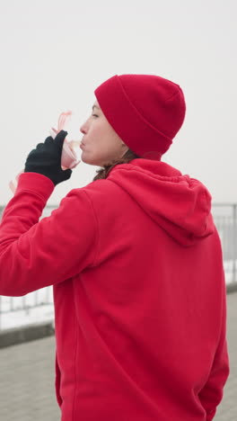 lady in red hoodie and beanie sipping water from pink bottle outdoors in serene winter setting with snow, iron railing, and distant misty bridge in background, peaceful atmosphere