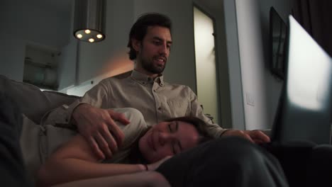 close up of a happy brunette girl in a beige t-shirt sleeping on the lap of her boyfriend who is working on a gray laptop in the evening remotely at home in a modern apartment