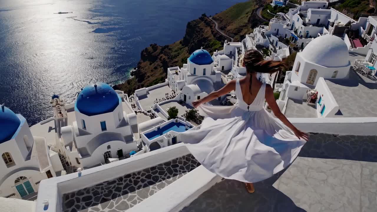 Aerial video captures a woman in a flowing white dress overlooking Santorini's iconic blue-domed