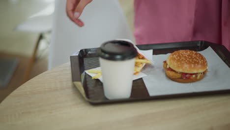 hand view of a woman in pink dress carrying a tray containing burger, fries, and coffee on a wooden table, she sits down and takes a potato chip, with a blurred background of the restaurant