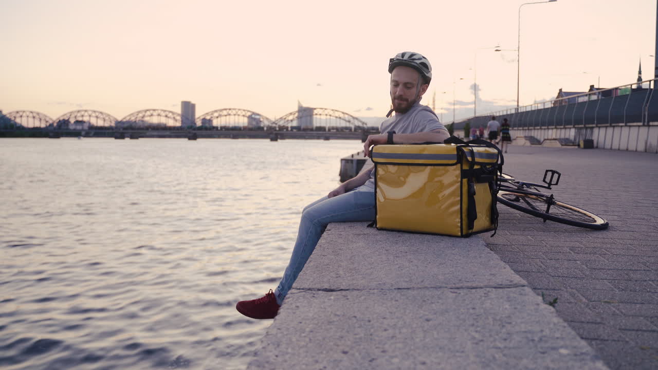 el repartidor de alimentos descansa junto a su mochila térmica al atardecer en la orilla de un río y echa un vistazo a su reloj inteligente