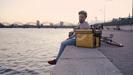 food delivery man rests next to his thermal backpack at sunset on the shore of a river and takes a look at his smartwatch