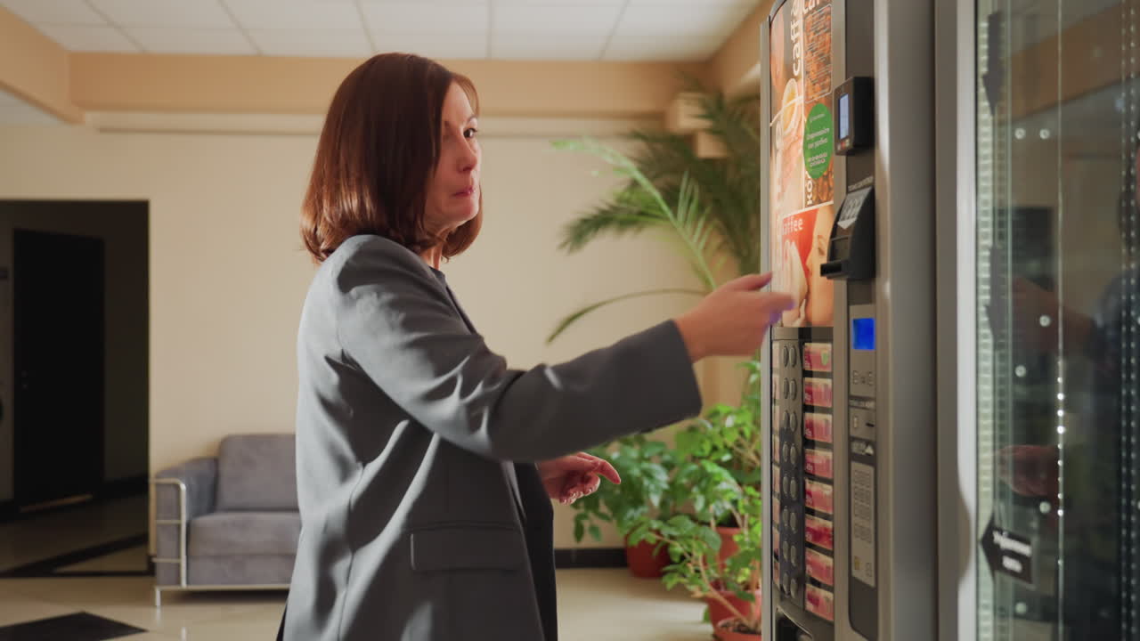 Mulher vestindo roupa de trabalho interagindo com máquina de venda automática no saguão de escritório moderno com sofá e plantas verdes no plano de fundo, selecionando bebida ou lanche para um rápido refresco durante um breve intervalo