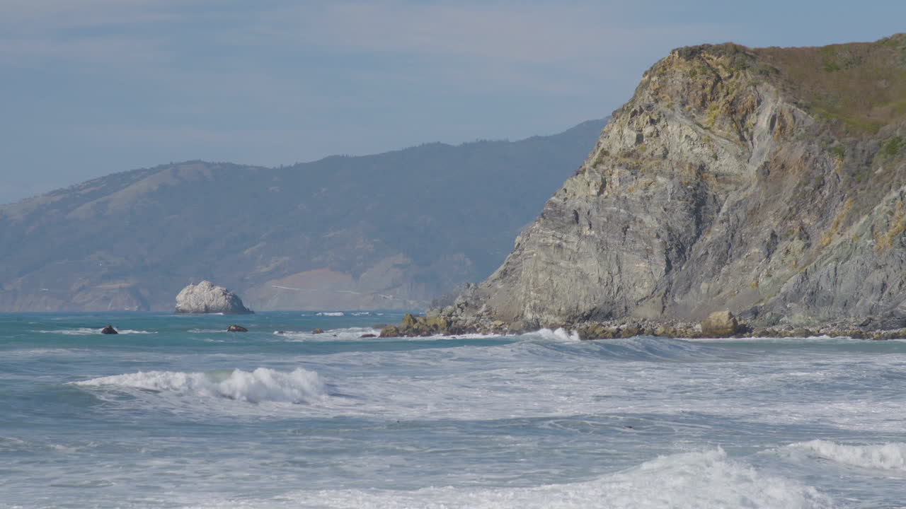 Inquadratura fissa di onde che si infrangono contro il fianco di un'alta scogliera, a Big Sur, California
