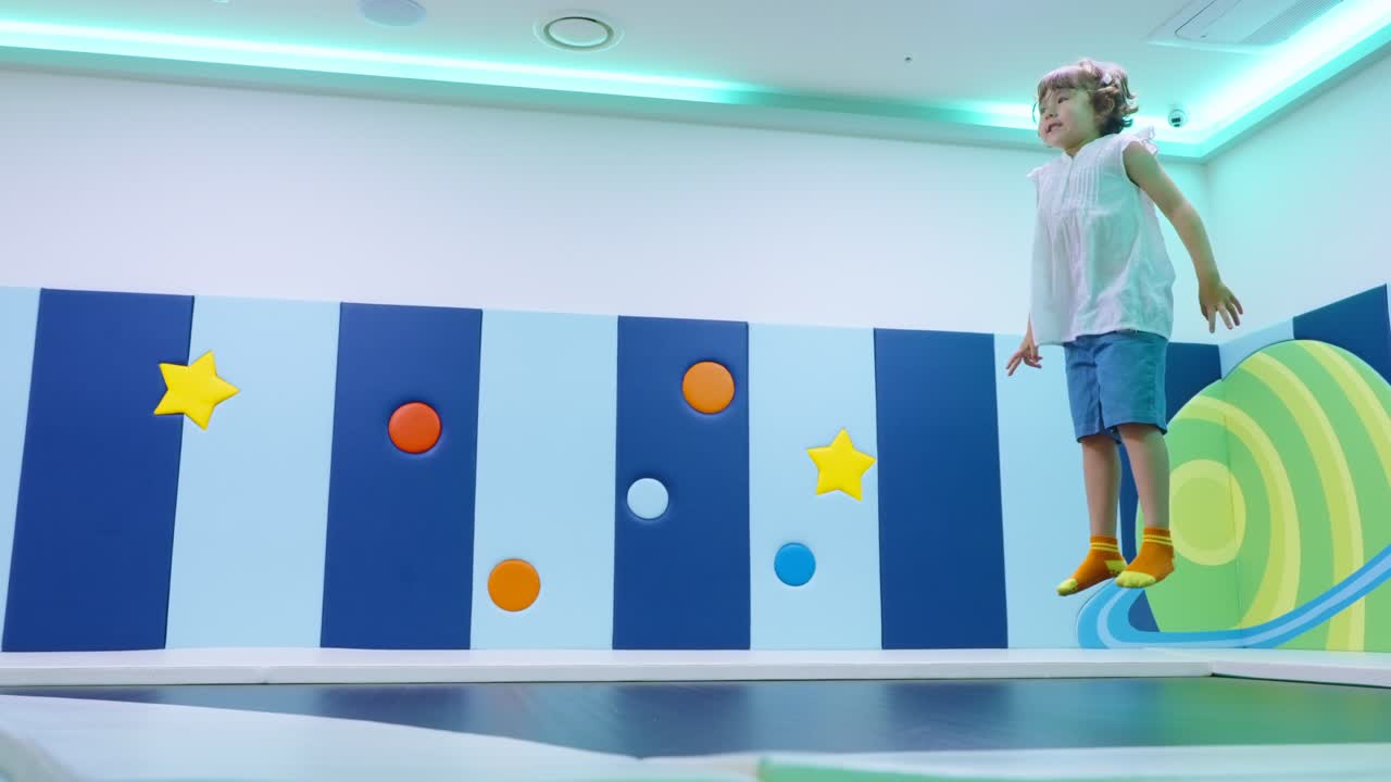 밝게 빛나는 우주 테마 방에서 5살 여자아이가 작은 실내 트램펄린 위에서 춤추고 포즈를 취하고 있어요