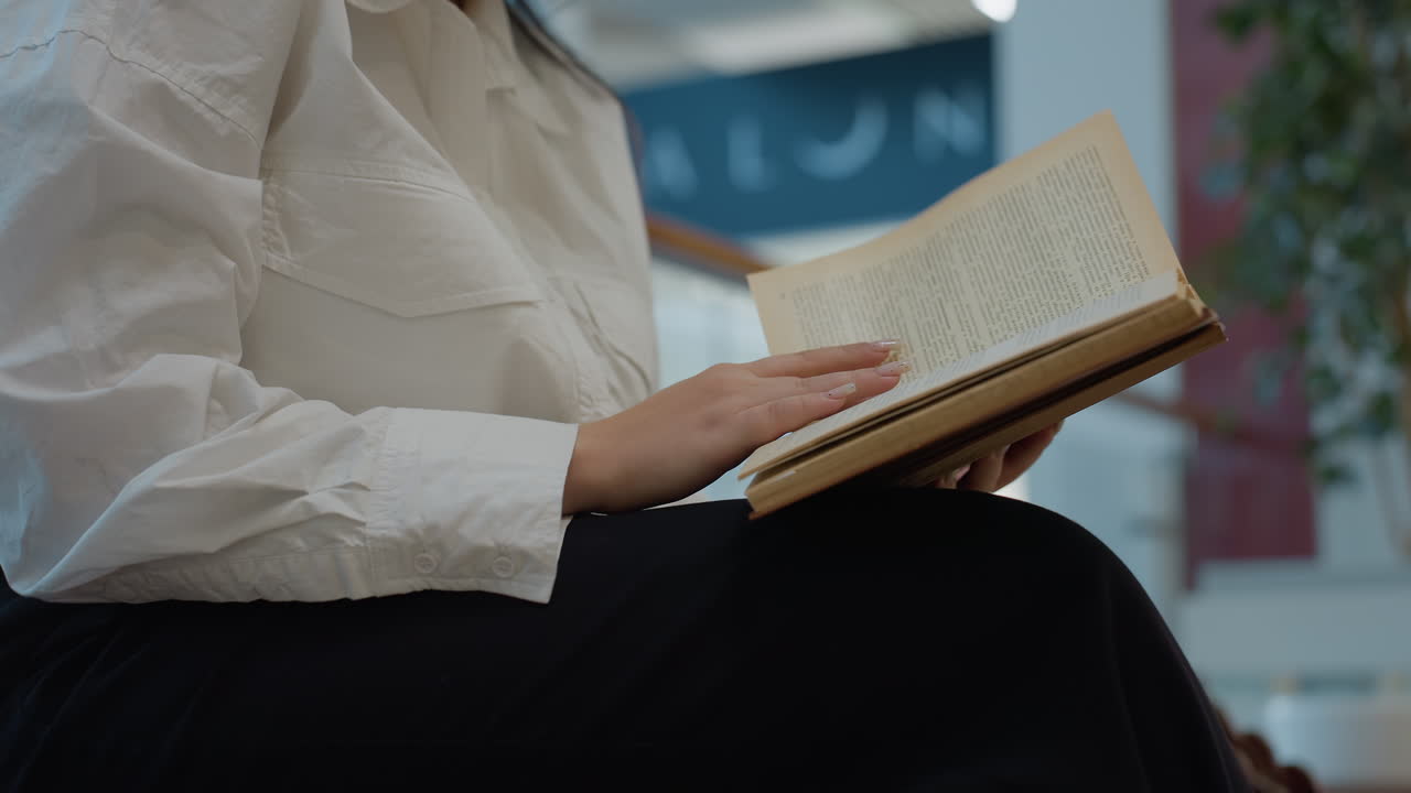 Thoughtful lady with long dark hair and glasses seated indoors carefully reads novel, gently flipping pages, surrounded by blurred floral displays, architectural glass panels and mall signage