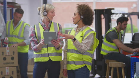 Dos-Trabajadoras-Que-Usan-Auriculares-En-Un-Almacén-De-Distribución-Logística-Usando-Una-Tableta-Digital