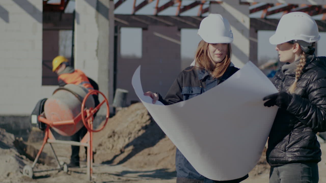 Young female contractor and architect wearing hardhats discussing over blueprint at incomplete construction site