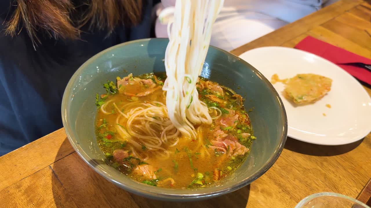 A person lifts rice noodles from a flavorful beef pho soup in a warmly lit restaurant setting