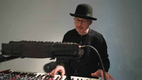 a keyboardist wearing a black hat, glasses, and a black outfit sits and plays a red sampler keyboard against a grey background, focusing intently on his performance