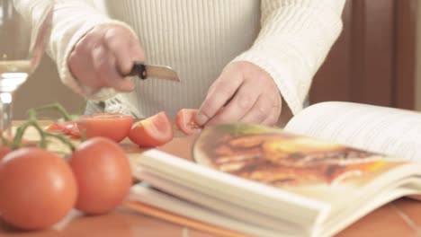 manos cortando tomates frescos en rama en la cocina con un libro de recetas de plano medio