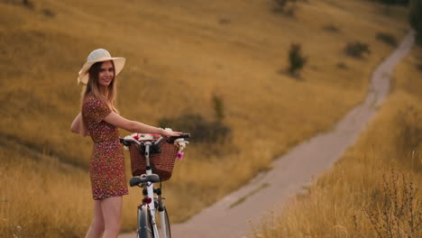 back kind of happy blonde girl in dress and hat turns around and smiling cheerfully looks at the camera and flirts strolling around the field in summer with bike and flowers.