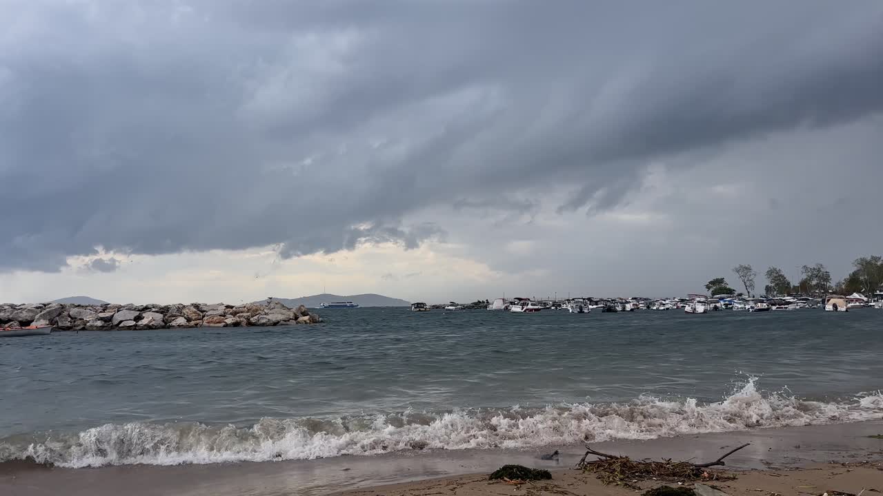 Dramatic dark storm clouds gather over a rough sea. Waves crash on the shore near a stone breakwater and a marina with boats on a gloomy autumn day