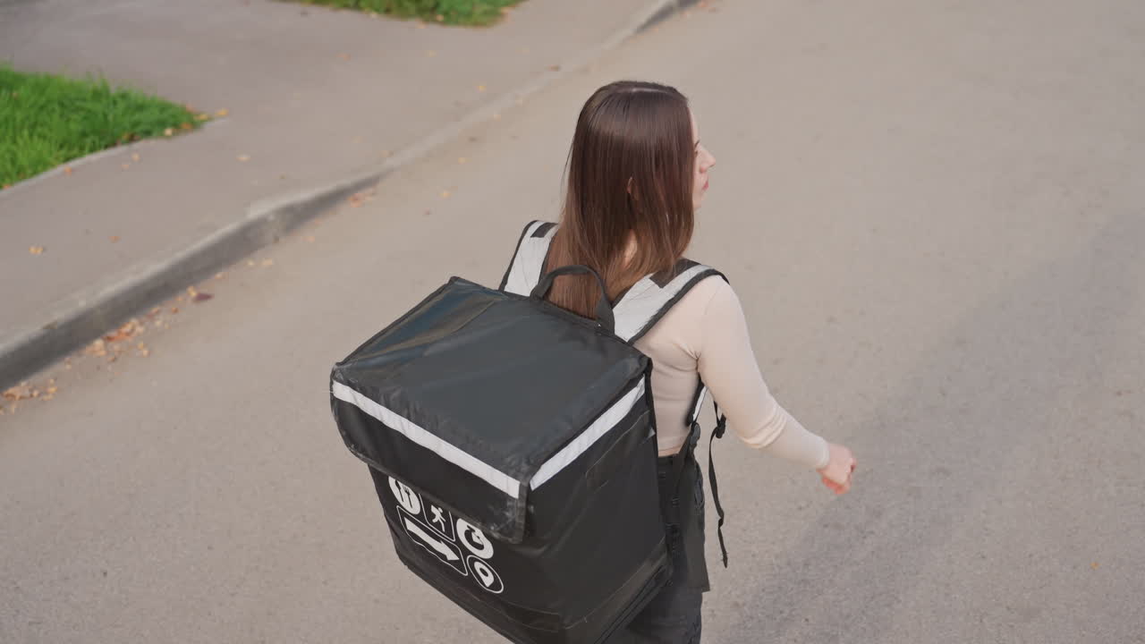 Mujer caucásica repartidora caminando por la calle con una mochila isotérmica grande, consultando su smartphone mientras se desplaza por una acera de las afueras cerca de un parque, tiras reflectantes en la mochila, chaqueta informal, pelo largo