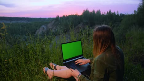 young girl works on a laptop with a green screen on top of a mountain.